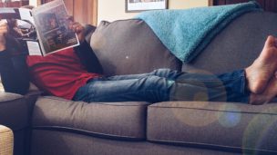 Man lays across couch with legs kicked up reading